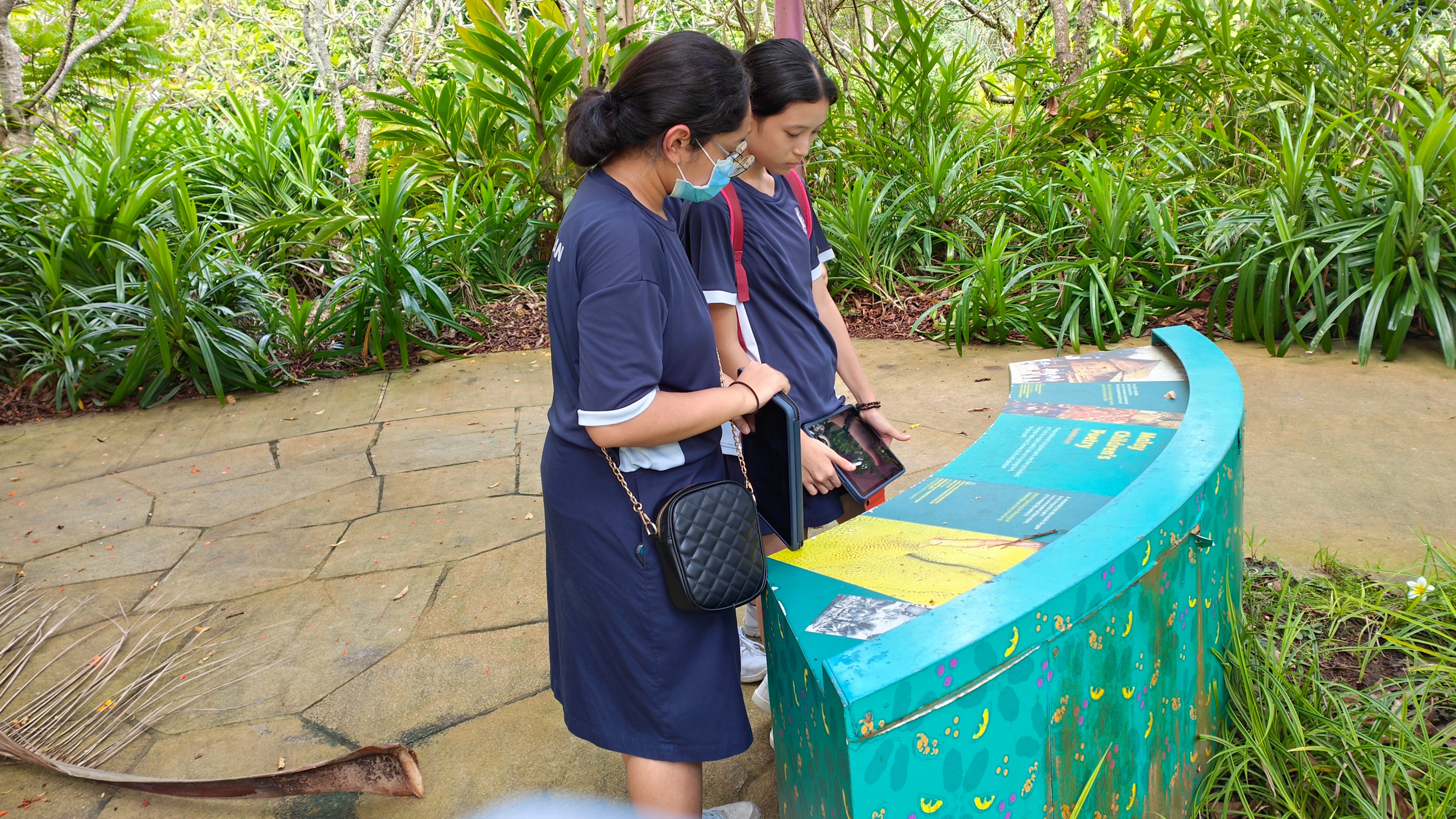 Two young girls in navy uniforms reading a teal curved outdoor information board surrounded by green plants.